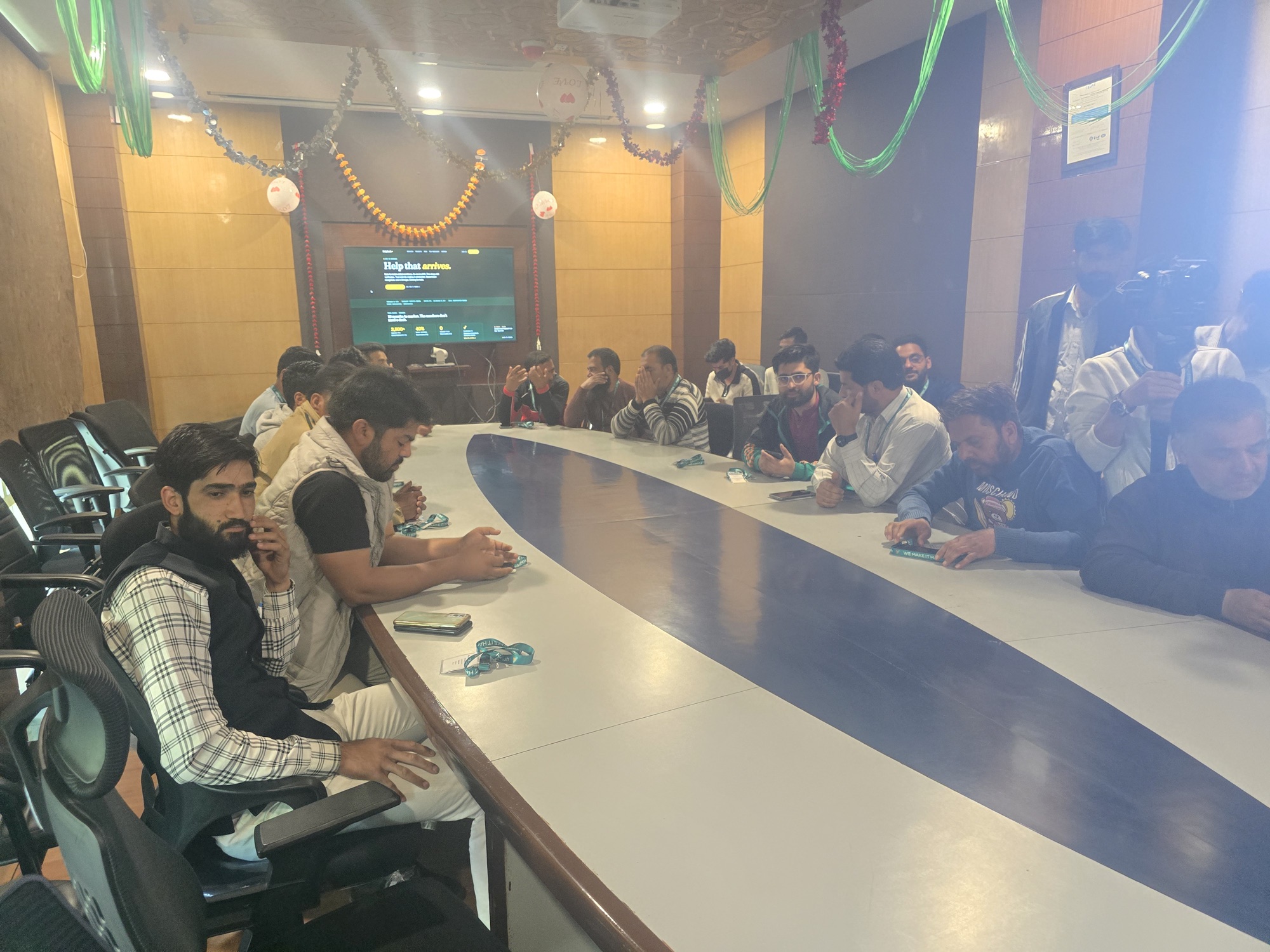 Founding Partners gathered around the long curved conference table at HelpRush's office in STPI Srinagar — the helprush.in 'Help that arrives.' homepage projected on the screen behind them, festive hall decoration overhead.