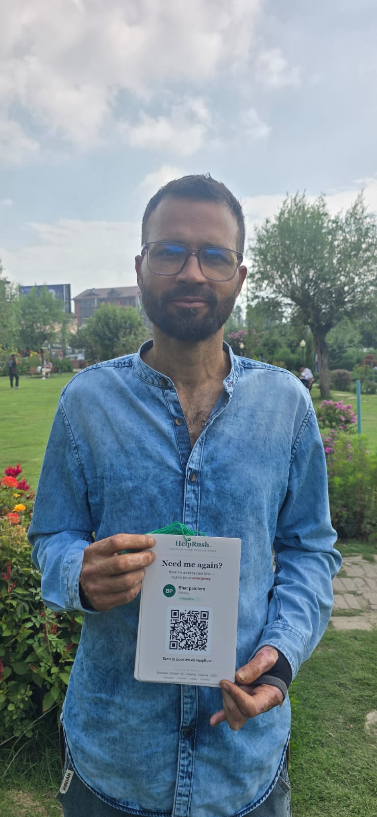 A painter in a denim shirt stands in a Srinagar park holding a HelpRush card at chest height. The card reads 'Need me again?' above his trade name, a verified-pro tick, and a large QR code. A loop of green string is threaded through the top.