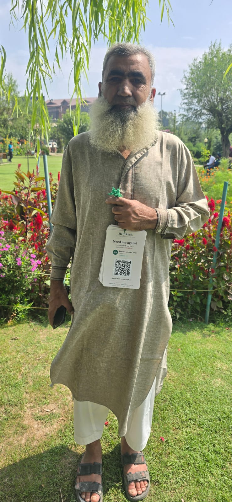 An older tradesman with a white beard, in a grey kurta, stands in a Srinagar park holding his HelpRush cards by the green string, looking directly at the camera.