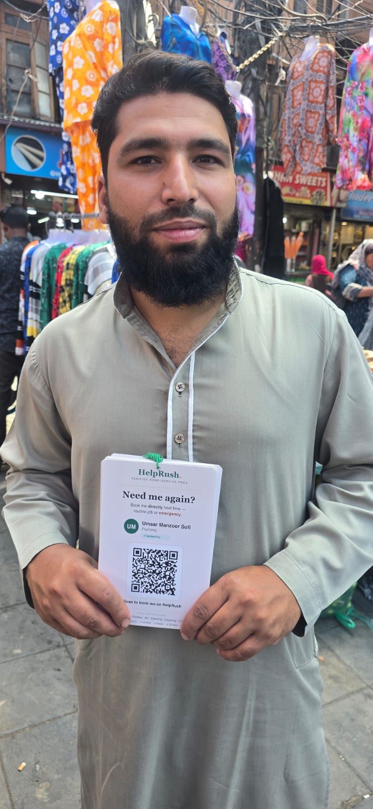A plumber stands in a busy Srinagar market lane, shopfronts hung with clothes behind him, holding a stack of his HelpRush cards. The top card shows his name, the word Plumbing, a verified-pro tick and a QR code.