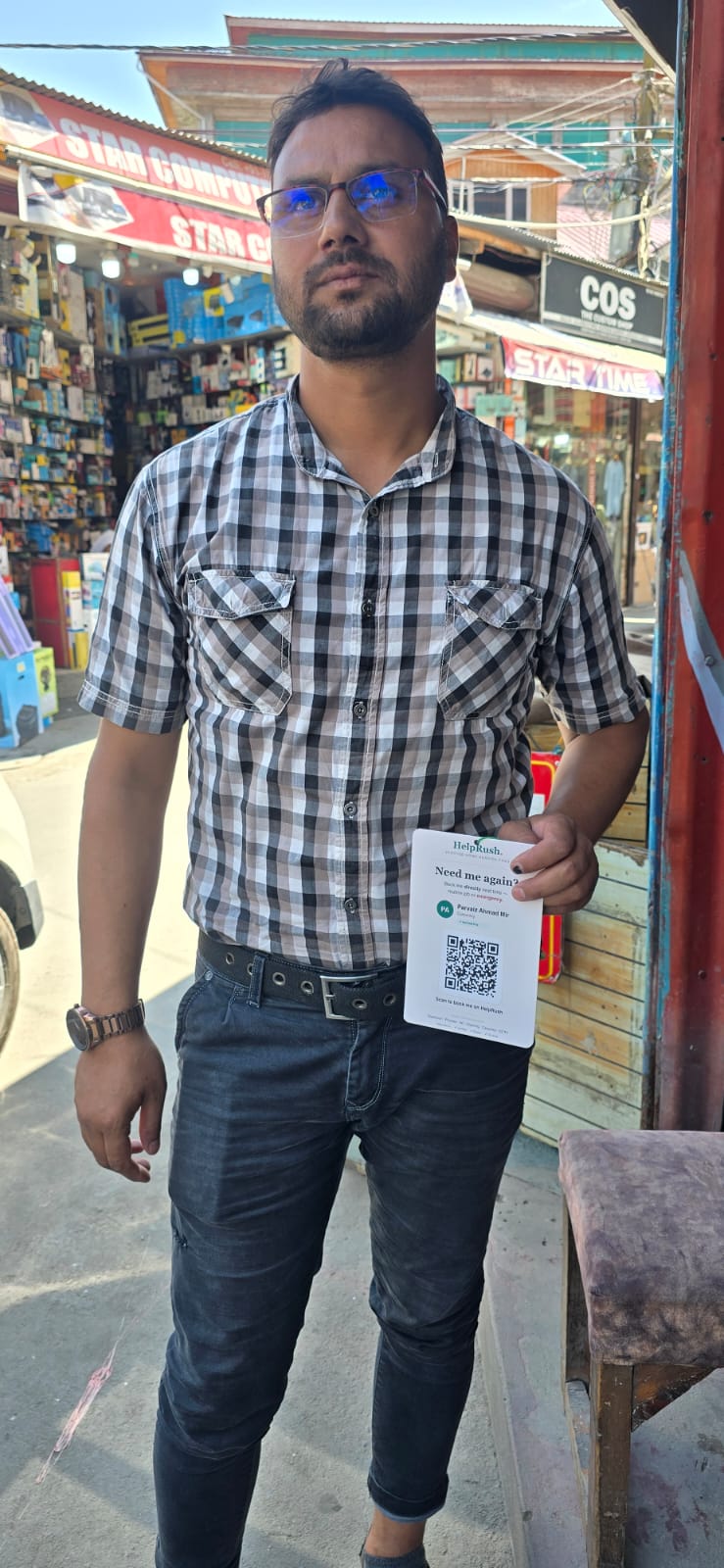 A tradesman in a checked shirt stands outside a computer-hardware shop in a Srinagar market, holding his HelpRush card with its QR code facing the camera.