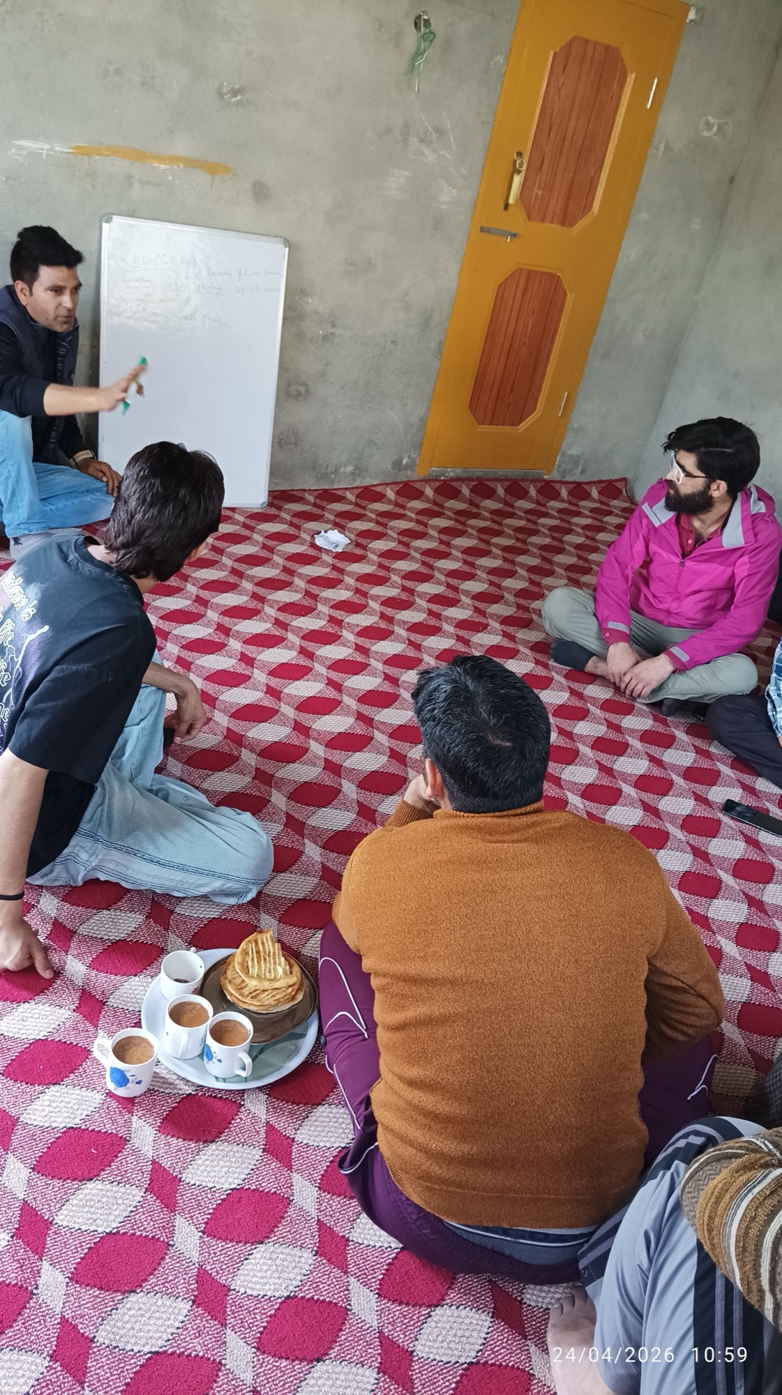 A small tray of mismatched cups holding kahwa sits on the patterned carpet beside a stack of chot kulcha; a partner in an orange sweater is seated foreground, the trainer in the corner writing on the whiteboard.