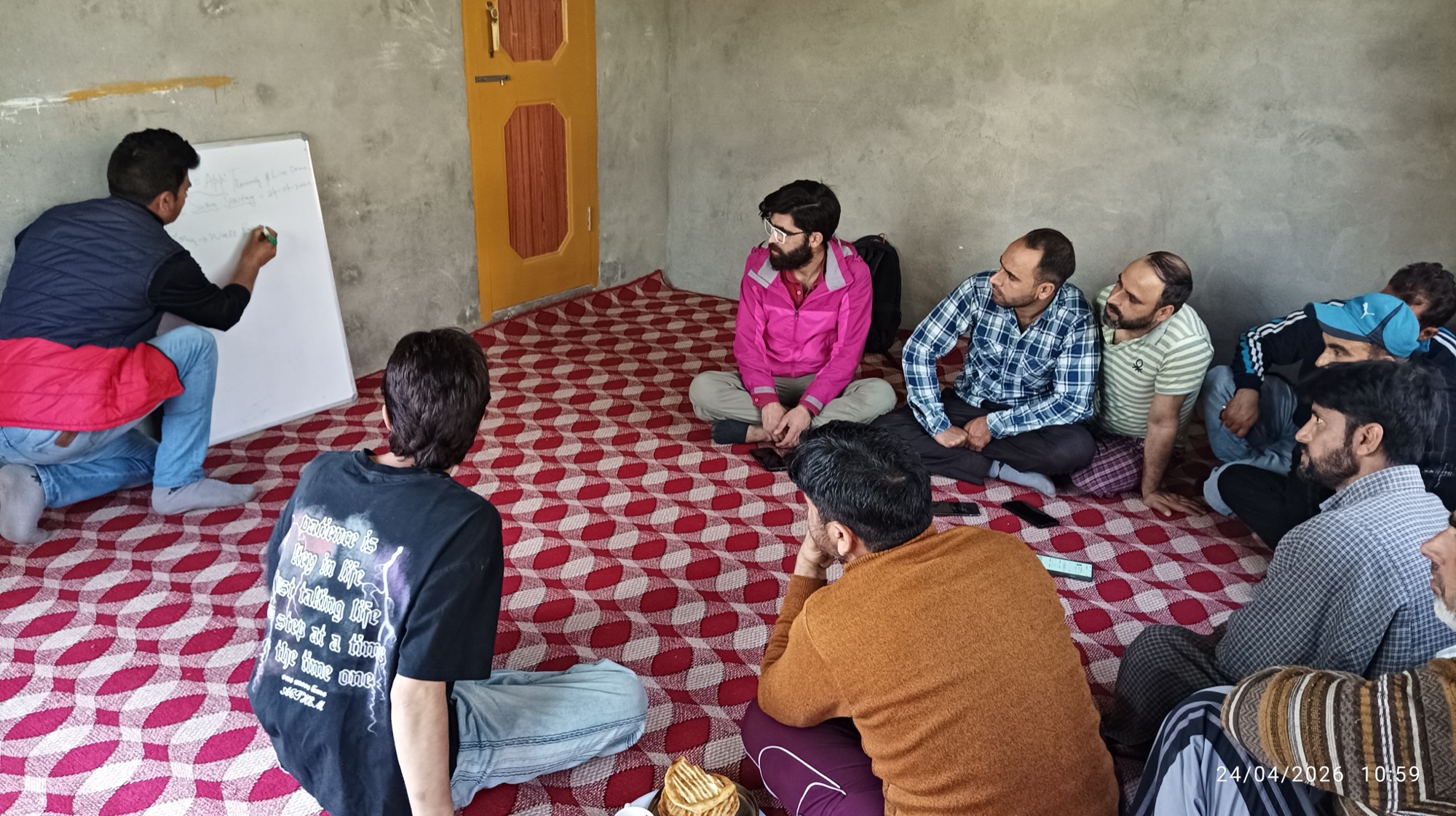 A HelpRush field-ops trainer writes on a whiteboard while seven partners sit cross-legged on a patterned carpet, one in a pink jacket facing the board attentively.