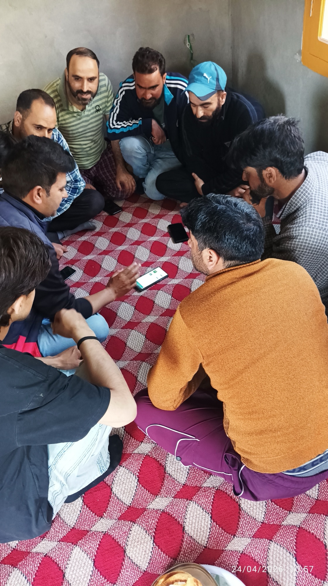 Eight men sit in a tight circle on a red-and-cream patterned carpet, attention focused on a single phone in the centre of the floor — a colleague in an orange sweater explains the screen.