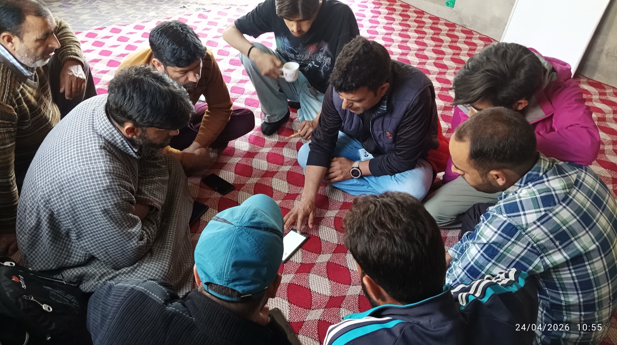 Close circle of eight partners around a phone showing a HelpRush job offer; a tea cup is held in the middle of the frame, partners lean in, hands gesturing as a colleague demonstrates the flow.