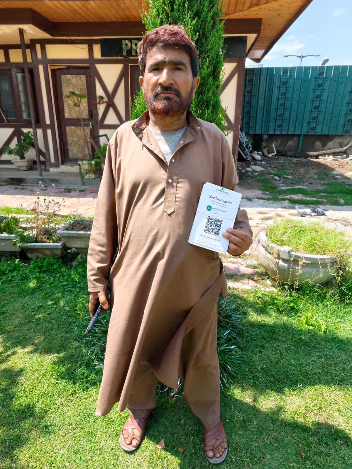 A tradesman in a brown kurta and leather sandals stands on the lawn outside the Pratap Park gatehouse in Srinagar, holding up his HelpRush 'Need me again?' card with its QR code, looking straight at the camera.
