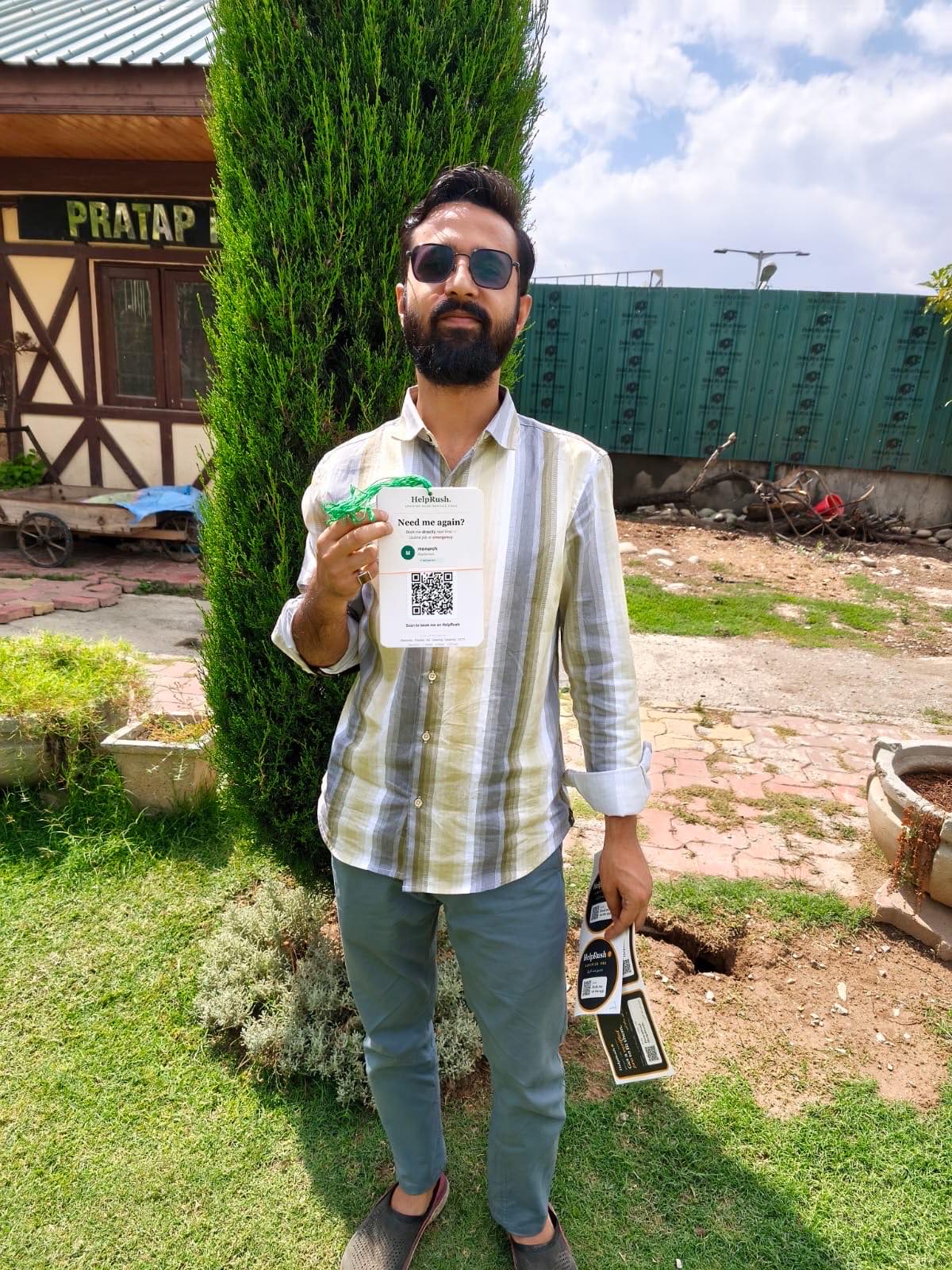 A young technician in a striped shirt and sunglasses stands in front of the Pratap Park gatehouse, holding up his new HelpRush card by its green string in one hand and a sheet of round Verified Pro stickers in the other.
