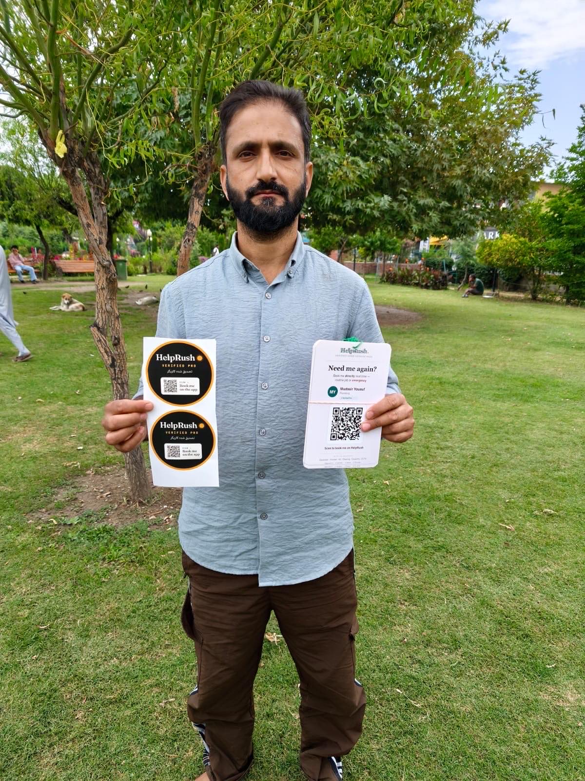 Mudasir Yousuf, a plumber, stands under a tree in Pratap Park holding up a sheet of two round HelpRush Verified Pro stickers in one hand and his personal 'Need me again?' booking card with its QR code in the other.