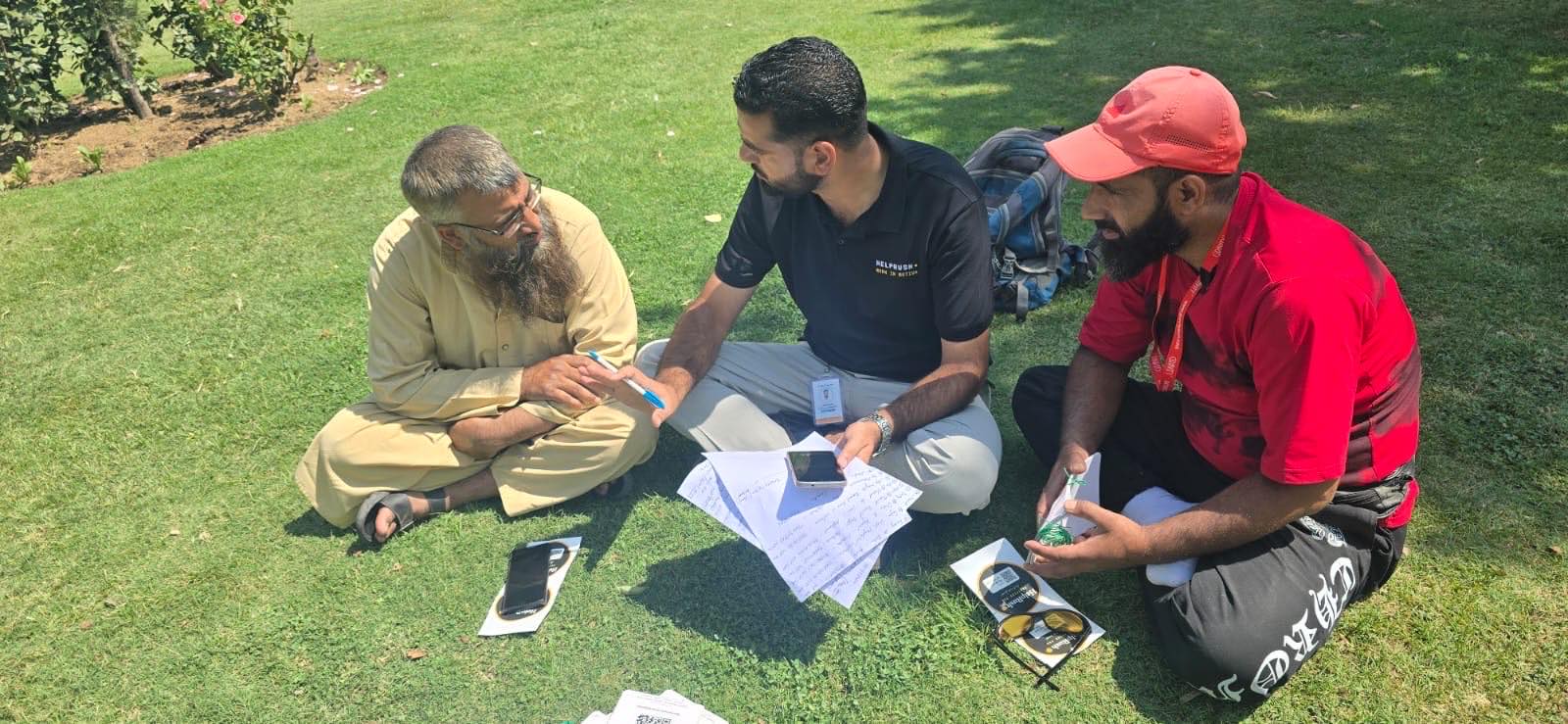 On the lawn at Pratap Park, a HelpRush field ops member in a black polo sits between two tradesmen, guiding the older one through the app on his phone. A sheet of round black Verified Pro stickers and a green-string card lie on the grass beside the man in the red cap.