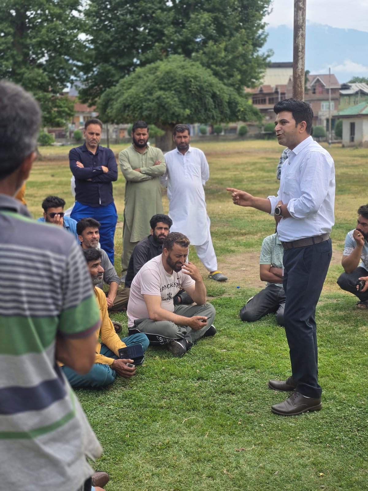 Showkat Kashkari, in a white shirt, stands on a park lawn in Soura addressing a group of blue-collar workers — some seated cross-legged on the grass, others standing with arms folded, listening.