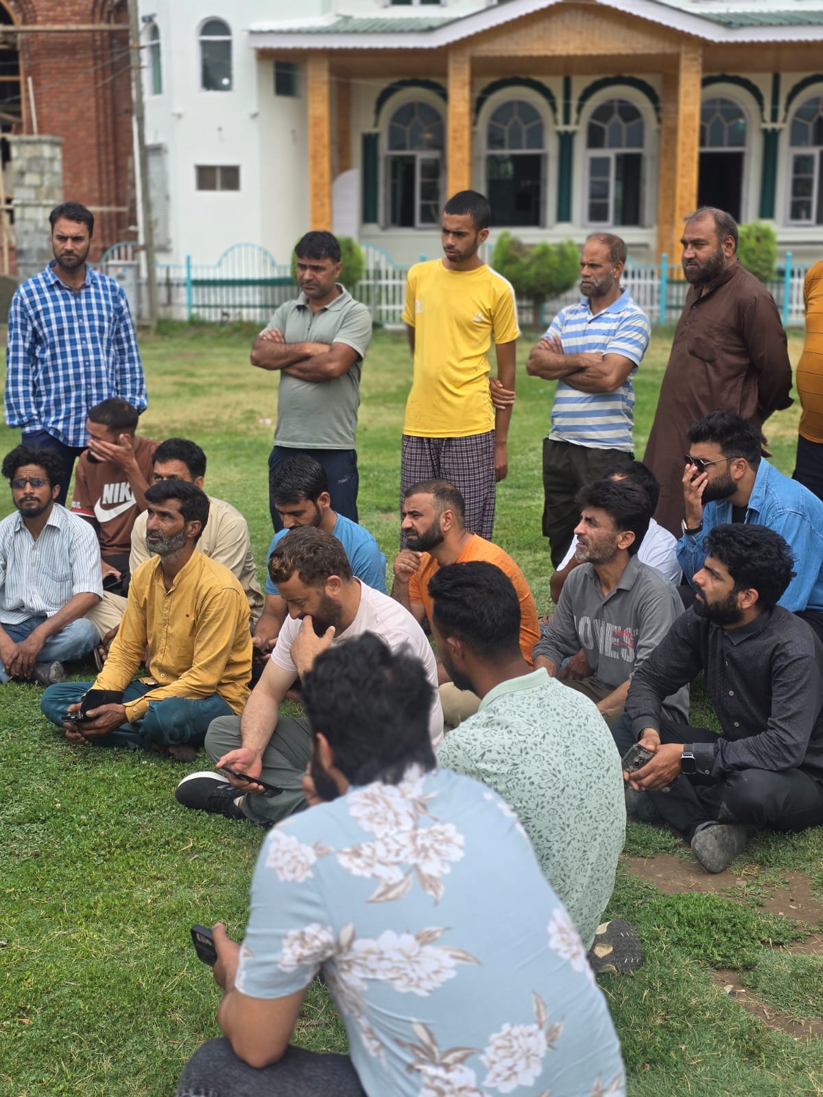 A few dozen workers sit on the grass in front of the masjid at Jenab Sahib — some with phones out, some listening with arms crossed — during the HelpRush onboarding drive.