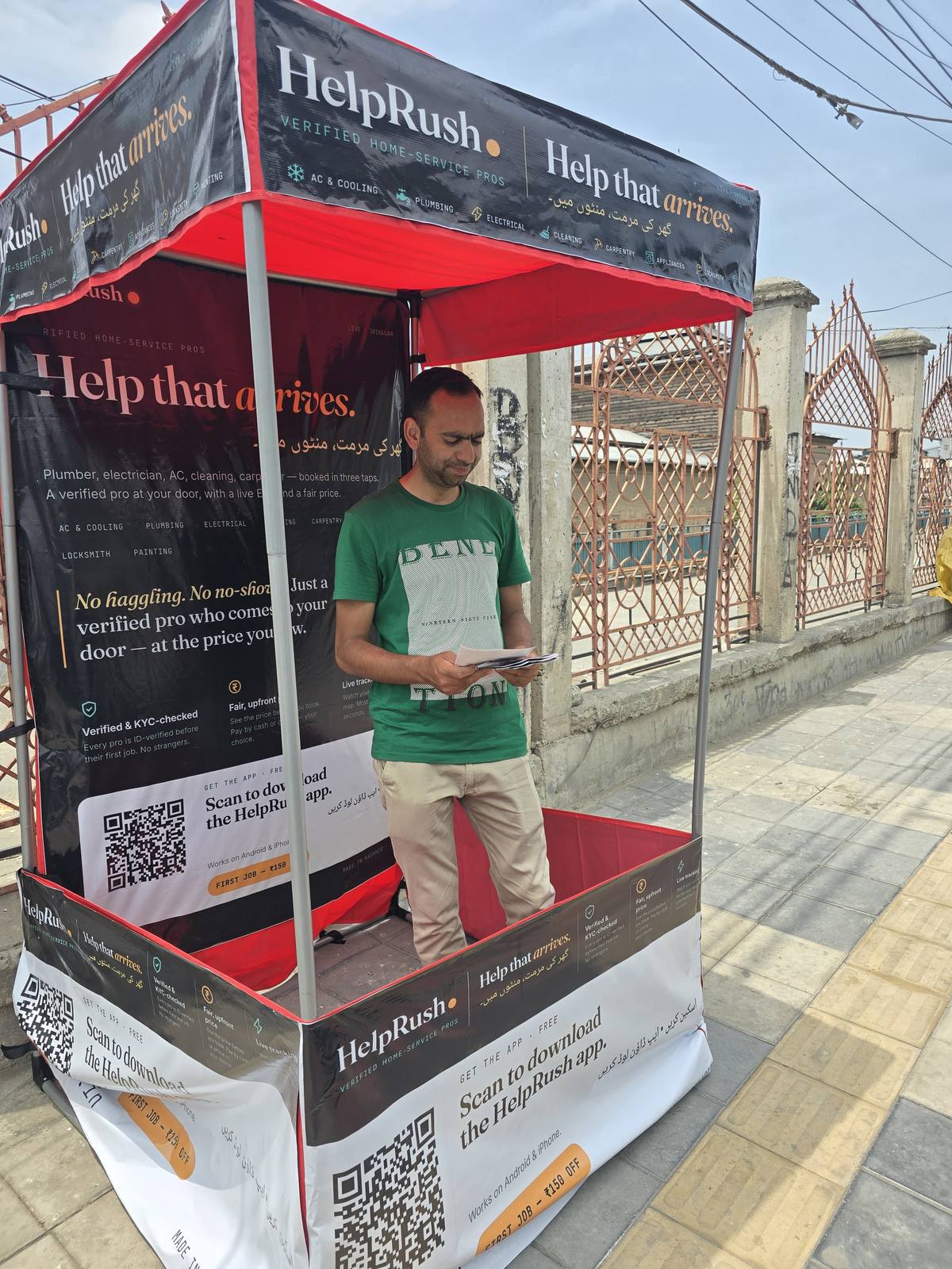 The HelpRush canopy seen from the side on the Nowhatta pavement, the masjid's iron gates and the street behind it, a field team member reviewing leaflets under the branded roof.