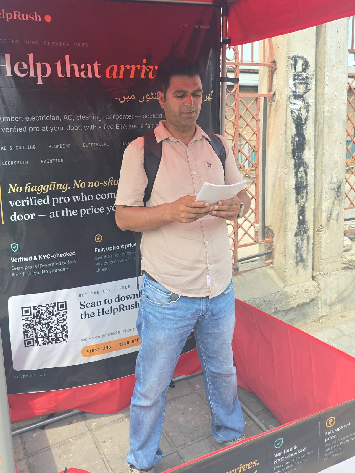 A HelpRush field team member with a backpack stands at the Nowhatta canopy reading through a stack of leaflets, in front of a banner listing services — AC and cooling, plumbing, electrical, locksmith, painting — with 'Scan to download the HelpRush app' and 'First job ₹150 off' printed below.