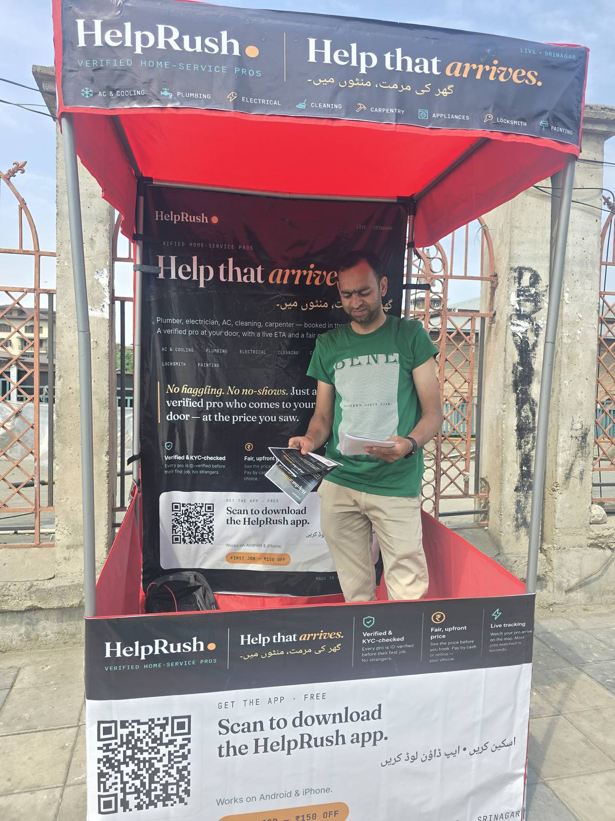 A HelpRush field team member stands inside a red-and-black HelpRush canopy pitched on the pavement outside the gates of Jamia Masjid, Nowhatta, holding pamphlets. The canopy reads 'Help that arrives' in English and Urdu, with a QR code to download the app.