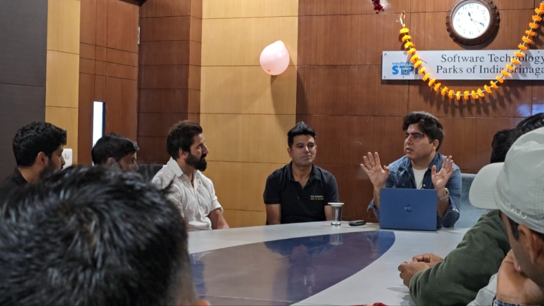 At the head of the oval table, three people face the room: a man in a white shirt, a HelpRush field ops member in a black HelpRush polo, and a presenter in a denim jacket mid-gesture behind an open laptop, under the STPI Srinagar sign and a marigold garland.