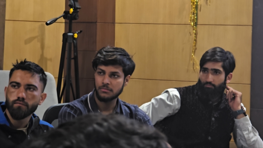 Three of the Super 30 listening from their chairs: a man in a blue jacket on the left, a young man in a dark shirt in the centre, and a bearded man in a black embroidered waistcoat on the right, chin on his hand. A camera tripod stands behind them.