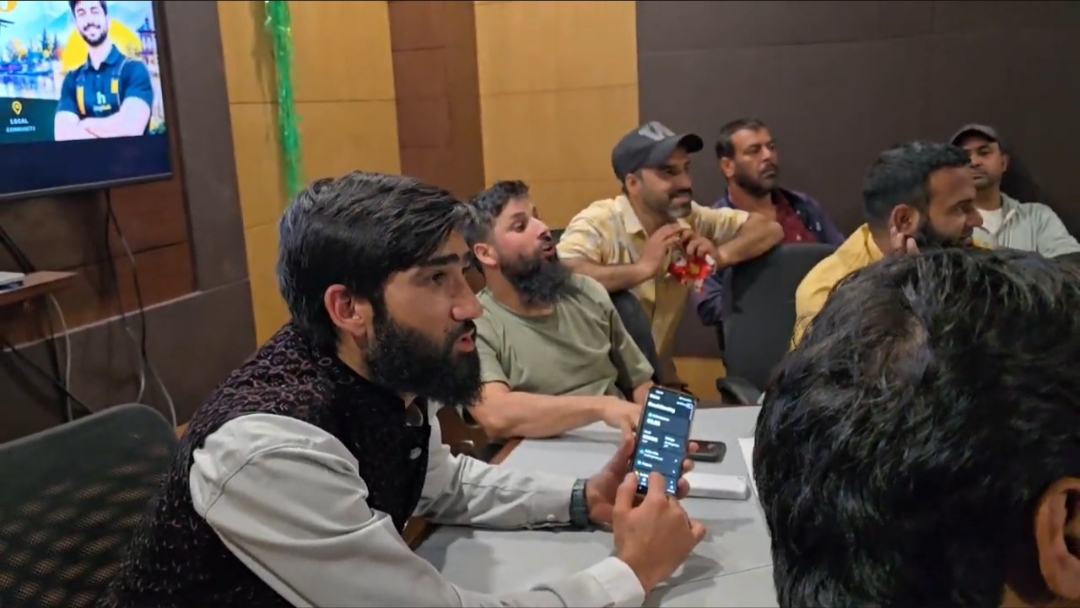A young tradesman in a black embroidered waistcoat over a white shirt holds up his phone, showing a HelpRush screen, while asking a question. Behind him four others lean in, one grinning, with a HelpRush partner poster on the television screen on the wall.