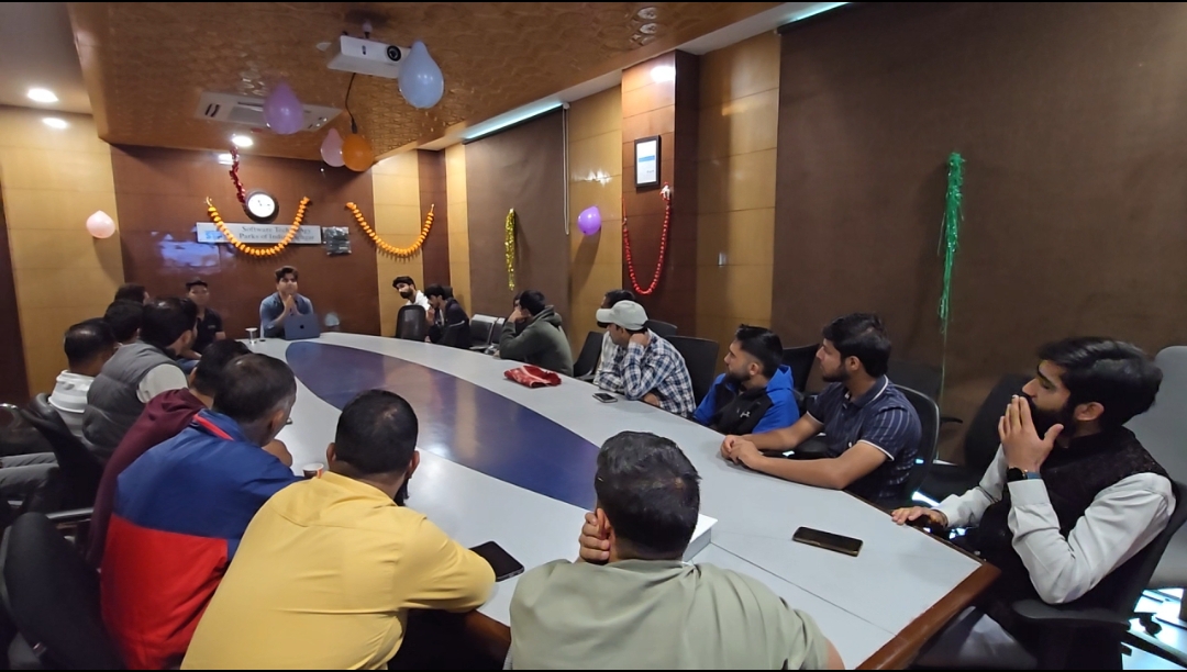 A wide view down the oval conference table from the near end. Tradesmen in jackets, kurtas and caps sit along both sides, phones face-down on the table, watching the HelpRush team member presenting at the far end under the STPI sign.